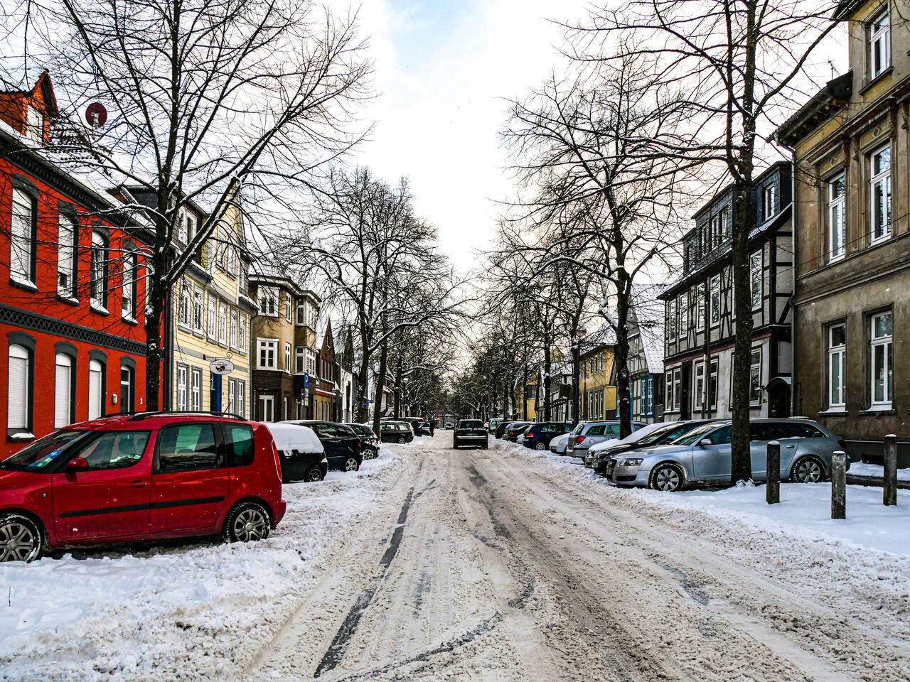 Schnebedeckte Straße mit parkenden Autos und einem sich entfernenden Fahrzeug.
